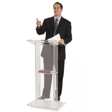 Man speaking behind the lectern with a book on the shelf, and a paper in his hand. 