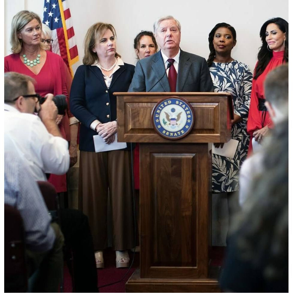 A man surrounded by people as he speaks behind an executive wood lectern